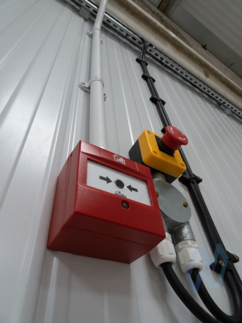 A fire alarm manual call point next to a machine emergency stop on the workshop wall