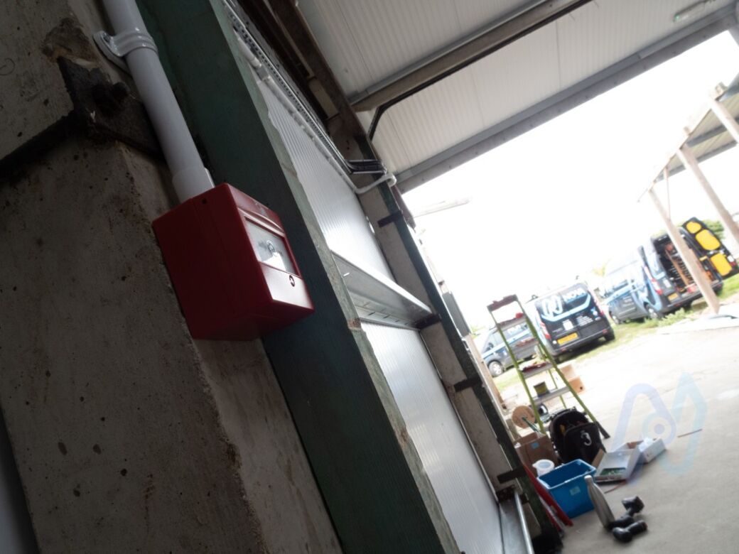 A manual call point beside the roller shutter door of a woodwork workshop in Ashford, with Nova vans outside