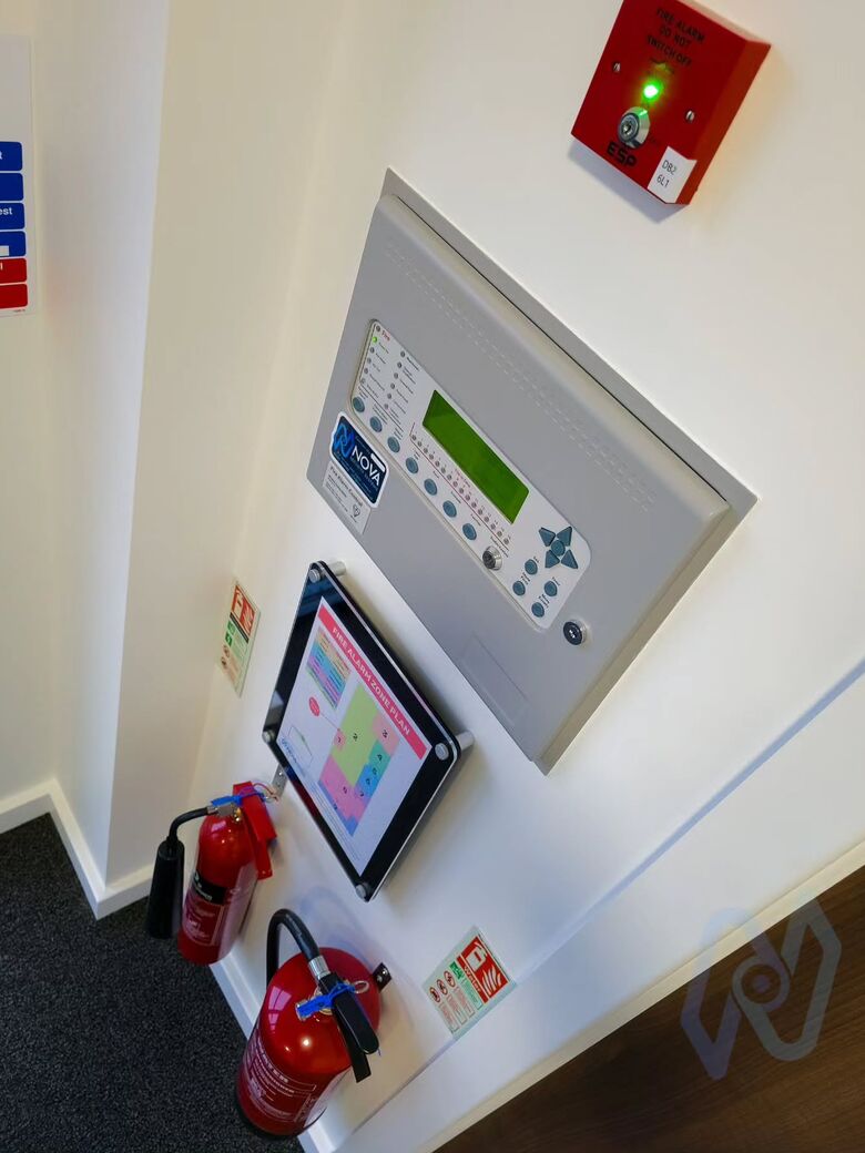 An addressable fire alarm panel with a framed zone plan, fire extinguishers and signs below