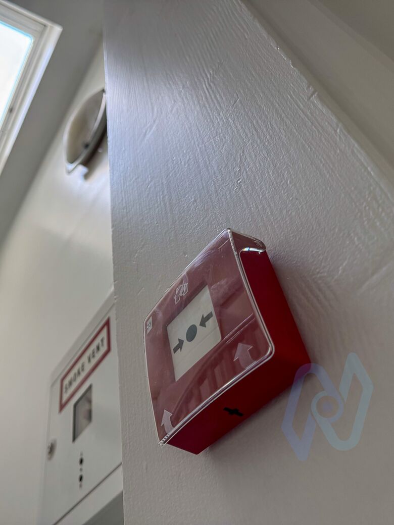 An Ajax EN54 manual call point with a clear protective cover, mounted on a wall corner